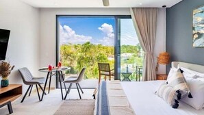 A modern hotel room with a large bed, a dining area with chairs, a ceiling fan, and a view of greenery outside. - C306 CPL, Radiante Studio Lagoon View-2 (Tulum)