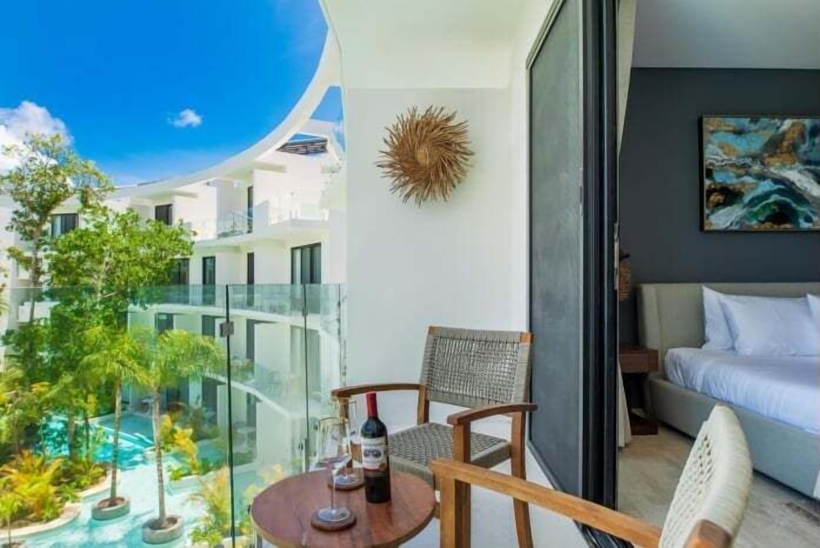 A balcony with a glass railing overlooking a pool and greenery, a table with a bottle of wine, and a bed with a painting on the wall.