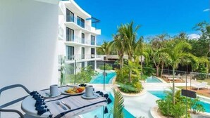 A balcony with a dining table set for two, overlooking a swimming pool area with palm trees and a modern building in the background. - D202 CPL, Gym|Spa|Coworking-2 (Tulum)