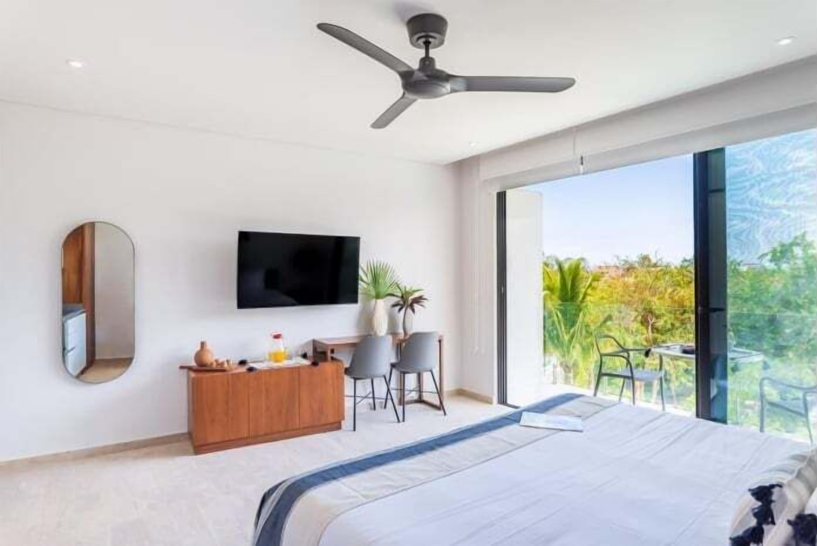 Une chambre moderne avec un grand lit, un ventilateur de plafond, une télévision, une commode en bois et un balcon agrémenté de plantes.