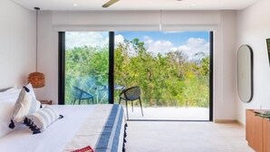 Et moderne soveværelse med en stor seng, loftventilator og en skydedør, der fører til en balkon med udsigt over grønne omgivelser.