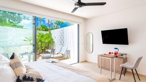 Ein modernes Hotelzimmer mit Bett, Schreibtisch, Sessel, Fernseher und Blick auf einen Garten durch eine Glasdoor.