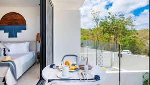 Una camera d'hotel moderna con un letto, un'area pranzo con tavolo apparecchiato per la colazione e un balcone con vista su alberi e cielo sereno.