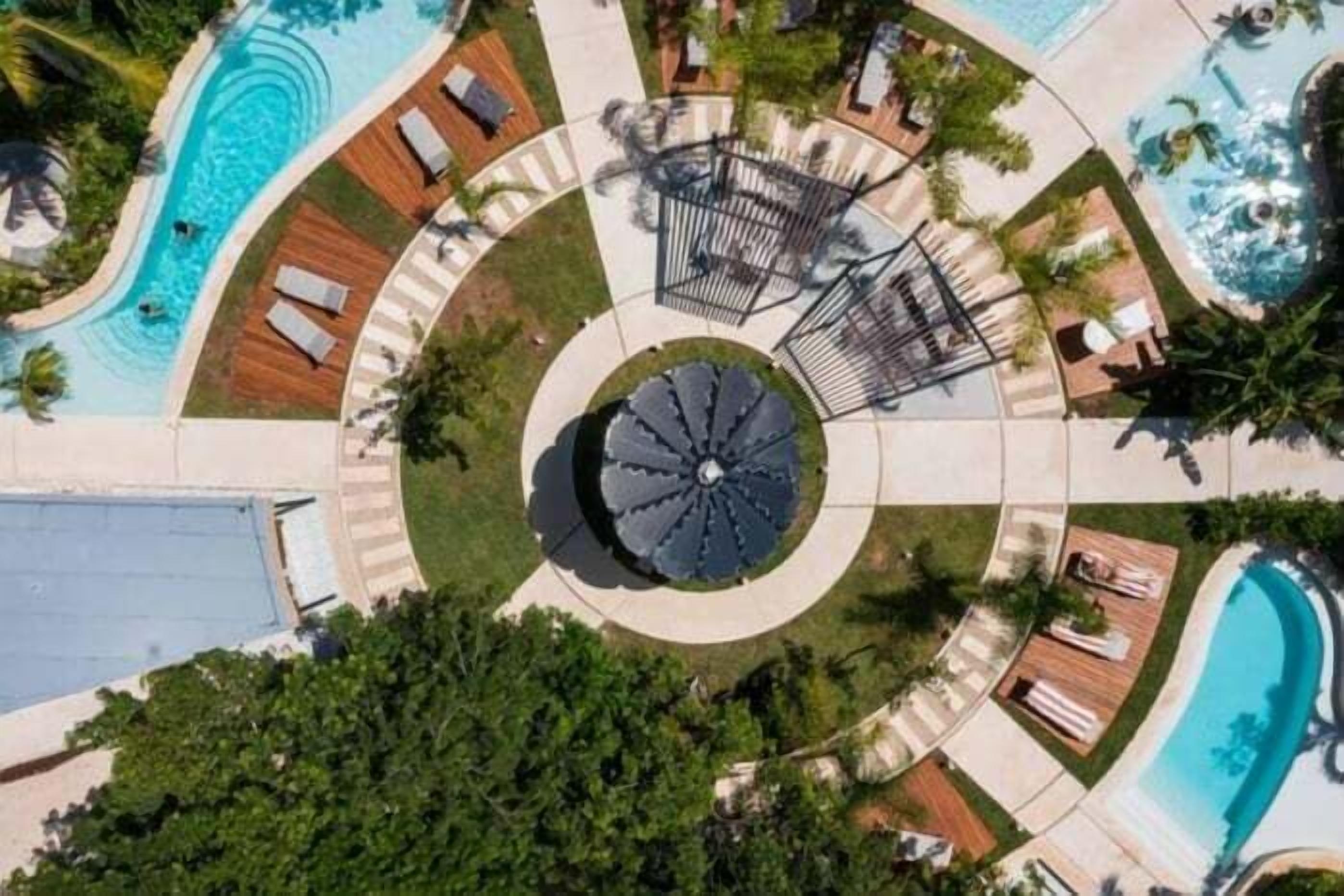A circular pool area with multiple lounge chairs and sunbeds, surrounded by a paved walkway and greenery.