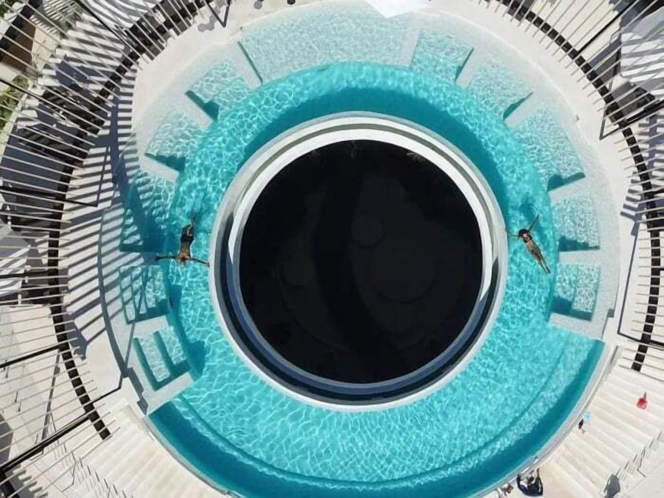 Una piscina a forma circolare con un’area centrale scura, circondata da un solarium bianco con ringhiere nere.