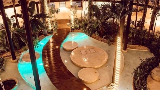 Jardín interior con sendero de madera, zonas de estar y plantas tropicales.
