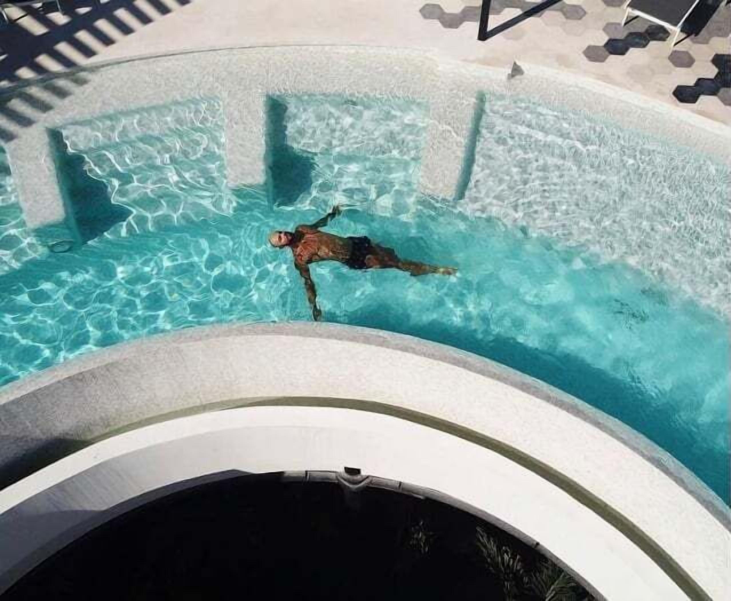 A person swimming in a clear pool with a tiled deck and lounge chairs in the background.