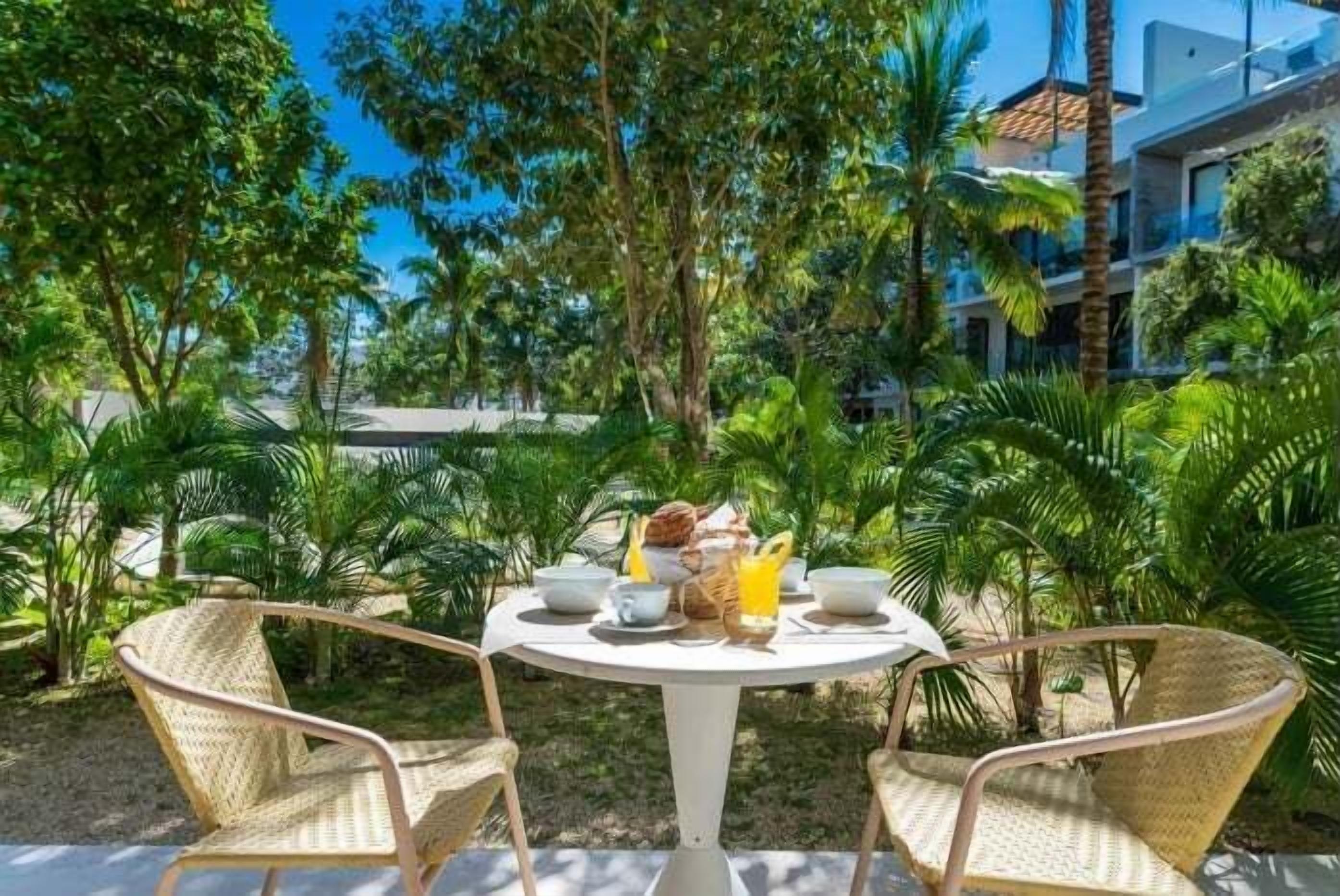 Ein runder Tisch mit zwei Korbstühlen, gedeckt mit Croissants, Orangensaft und Kaffeetassen, umgeben von üppiger Vegetation und einem modernen Gebäude im Hintergrund.