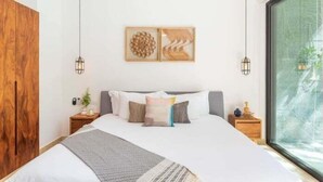 A modern bedroom with a large bed, a wooden wardrobe, a nightstand with a lamp, and a window with a view of greenery. - 104B PANOR, Modern Studio with 360° Pool-2 (Tulum)