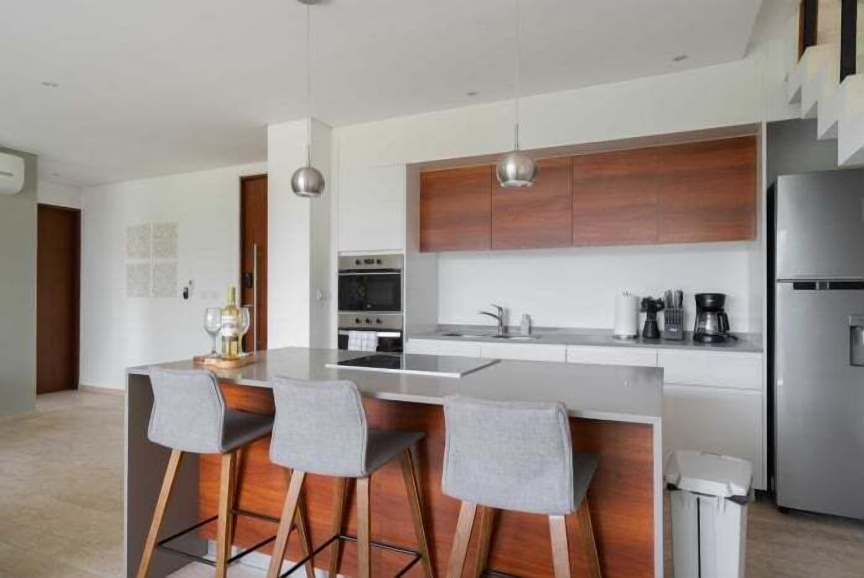 Una cocina moderna con una isla central, taburetes de bar, electrodomésticos de acero inoxidable y armarios de madera.