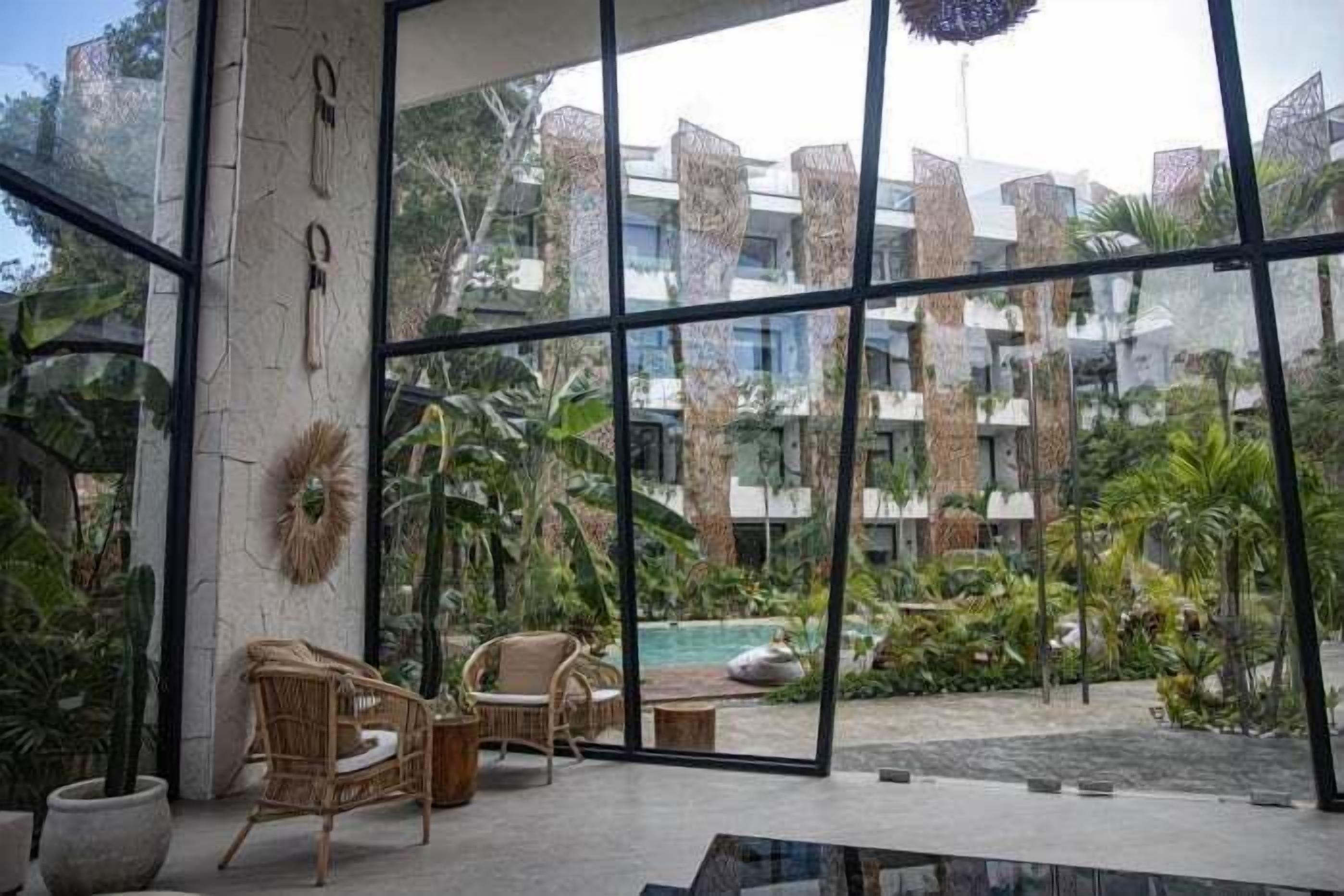 A modern hotel lobby with large glass windows, wicker furniture, and a view of a pool and greenery.