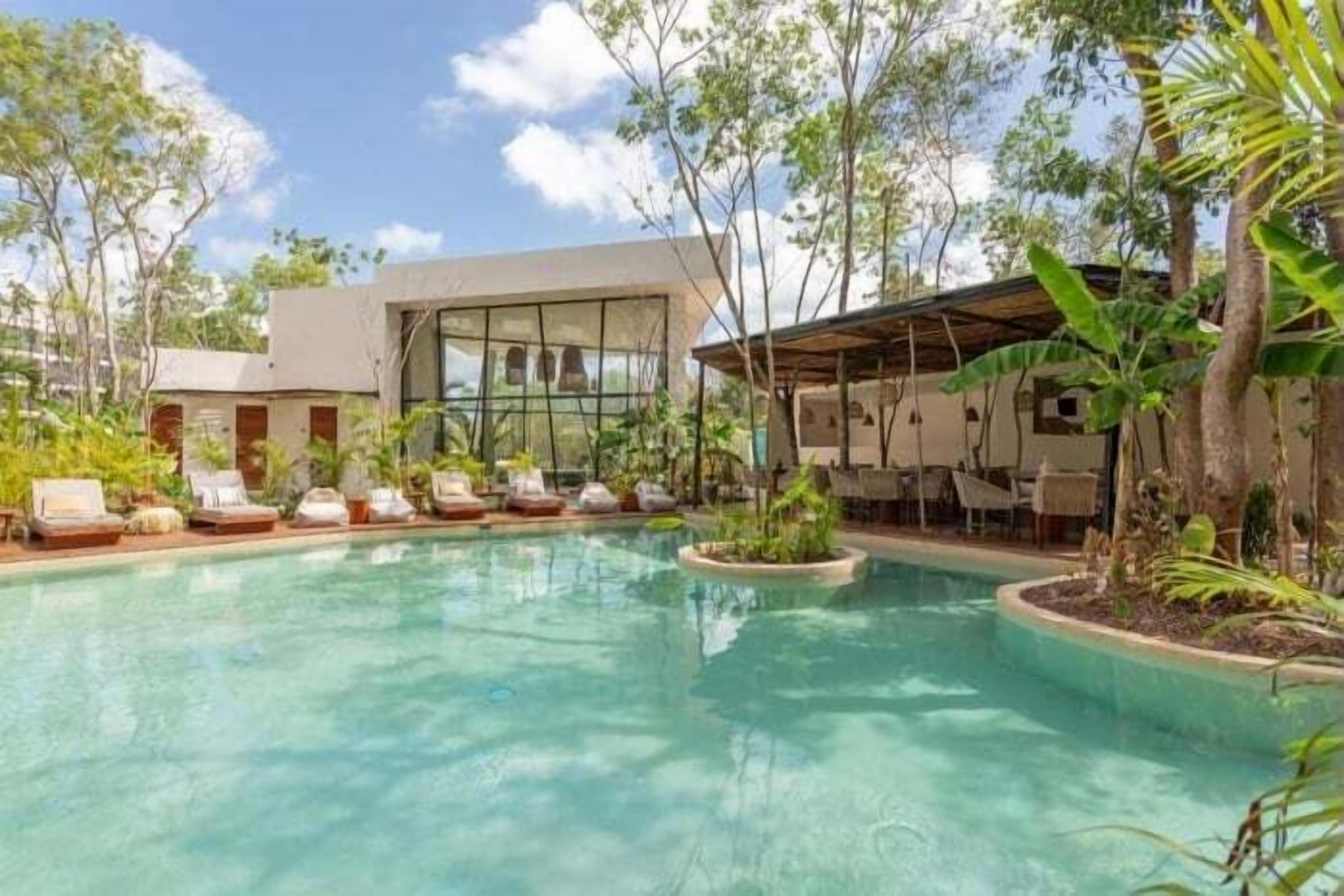 A pool area with lounge chairs and a covered seating area, surrounded by trees and a building with large windows.