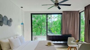 Een moderne hotelkamer met een groot bed, een plafondventilator, een zithoek met stoelen en een tafel, en uitzicht op groen.