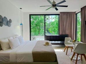 A modern hotel room with a large bed, a ceiling fan, a dining area with chairs and a table, and a view of greenery outside. - G204 CPT, Studio Serenity Escape-2 (Tulum)