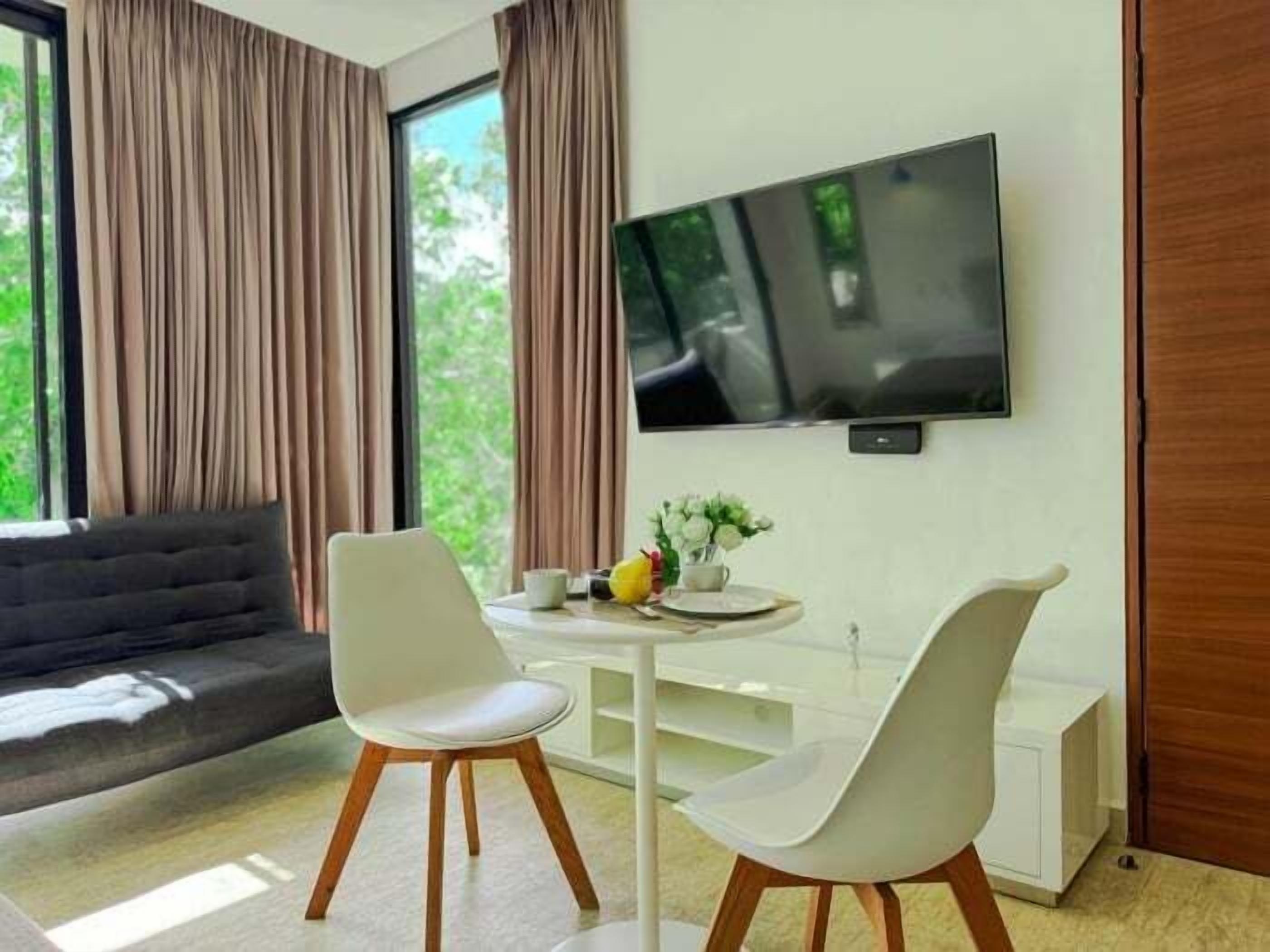 Een moderne woonkamer met een bank, een eettafel met twee stoelen, een flatscreen televisie aan de muur en een raam met gordijnen.