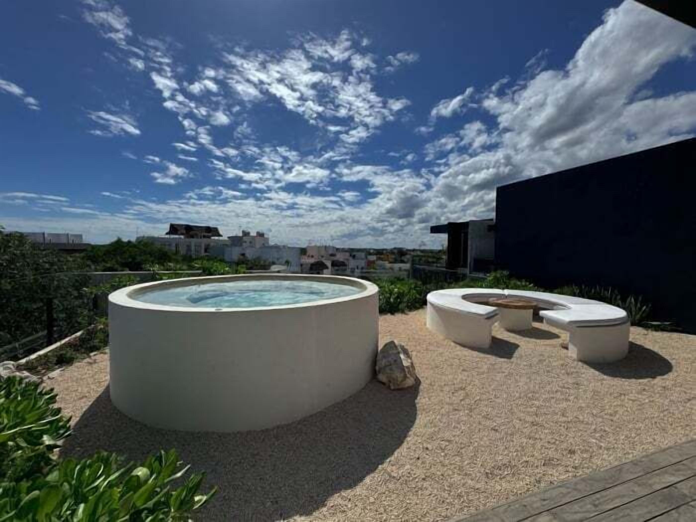 Et tagområde med swimmingpool, jacuzzi og siddepladser, omgivet af planter og en klar himmel.