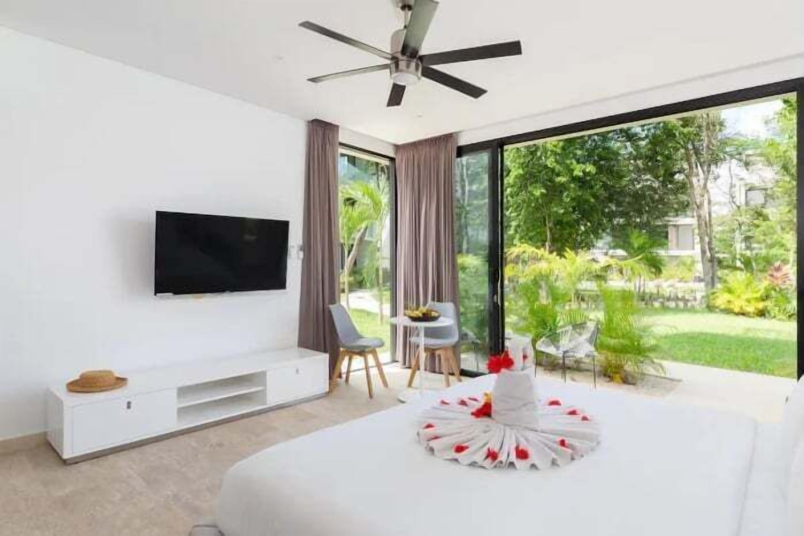Ein modernes Hotelzimmer mit einem großen Bett, einem Flachbildfernseher, einem Deckenventilator und Blick auf einen üppigen Garten.
