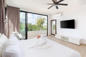 Ein modernes Hotelzimmer mit einem großen Bett, einem Flachbildfernseher, einem Deckenventilator und einem Balkon mit Blick auf Bäume.