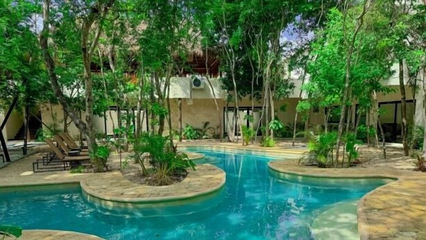 A pool with curved edges, surrounded by trees and plants, and a building in the background. - NAIA C19, Increible Studio Con Jacuzzi (Tulum)
