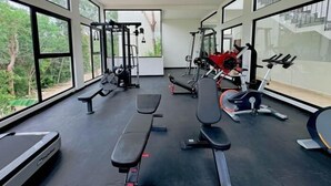 Ett gym med olika typer av träningsutrustning, stora fönster och utsikt över grönområden.