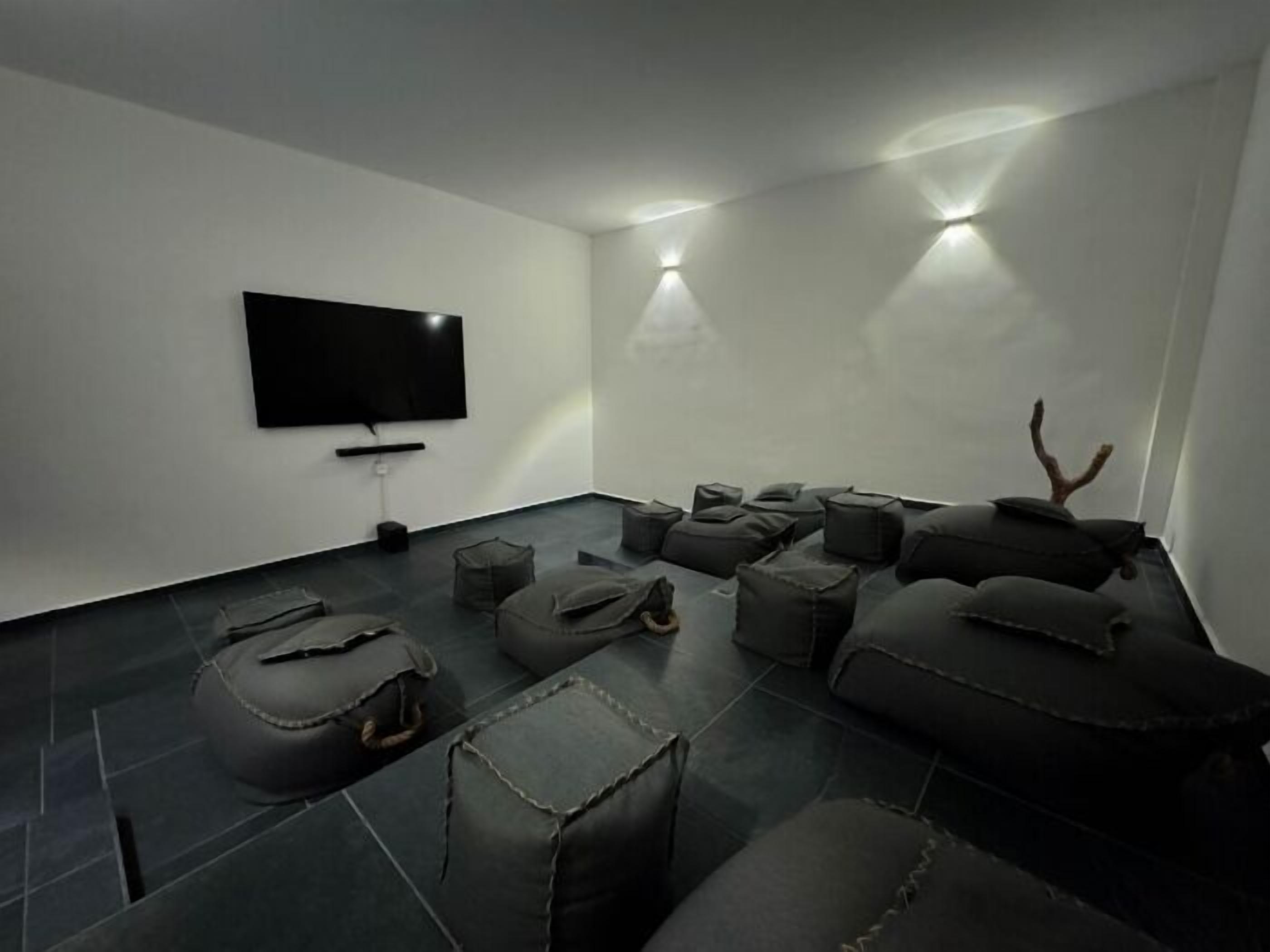 Ett modernt, minimalistiskt rum med en platt-TV som är monterad på väggen, en rad bekväma sittplatser och ett litet bord med en lampa.