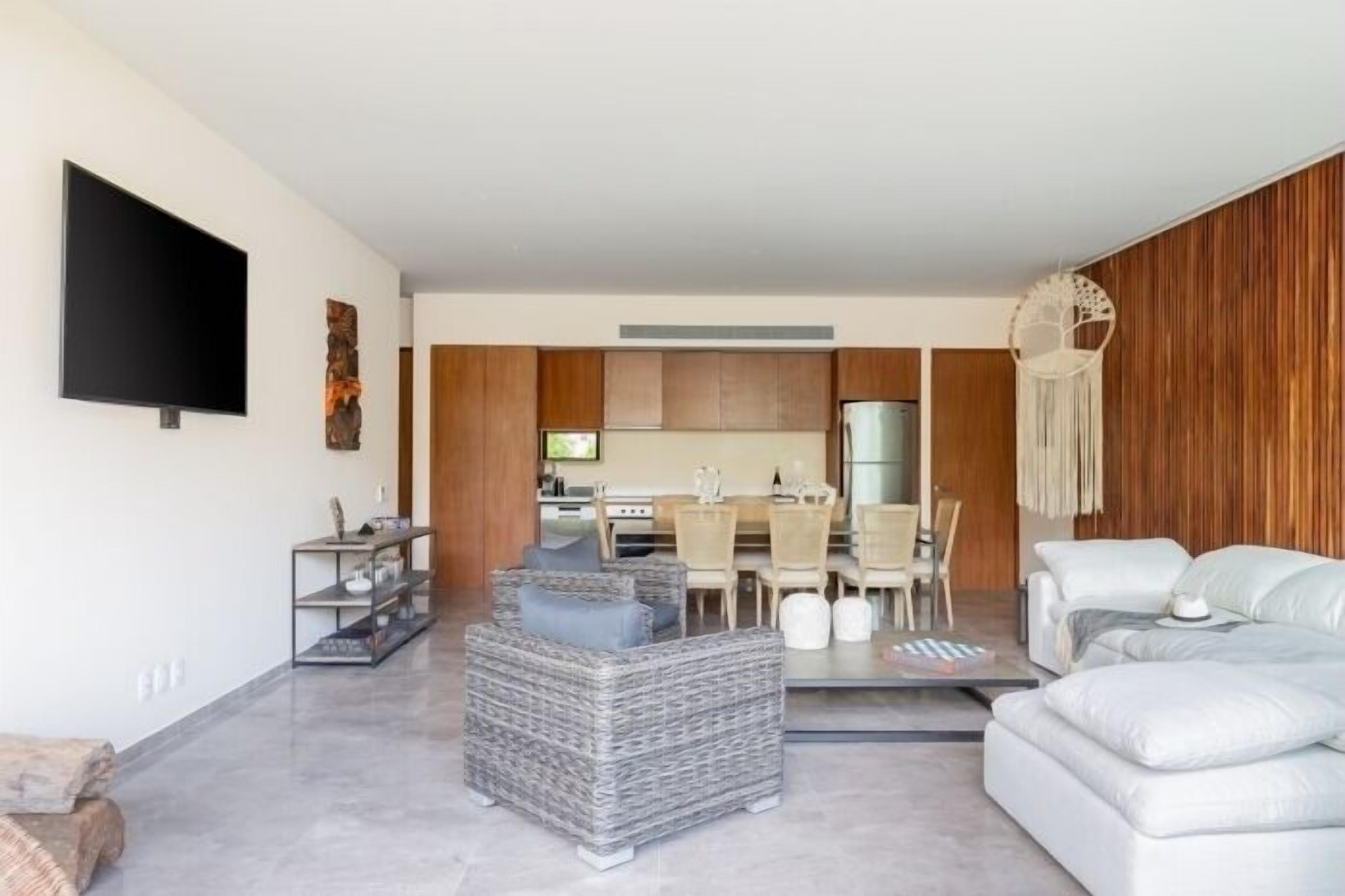 A modern living room with a grey wicker sofa, a white sofa, and a dining area with wooden cabinets and a TV.