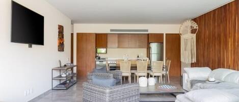 A modern living room with a grey wicker sofa, a white sofa, and a dining area with wooden cabinets and a TV.