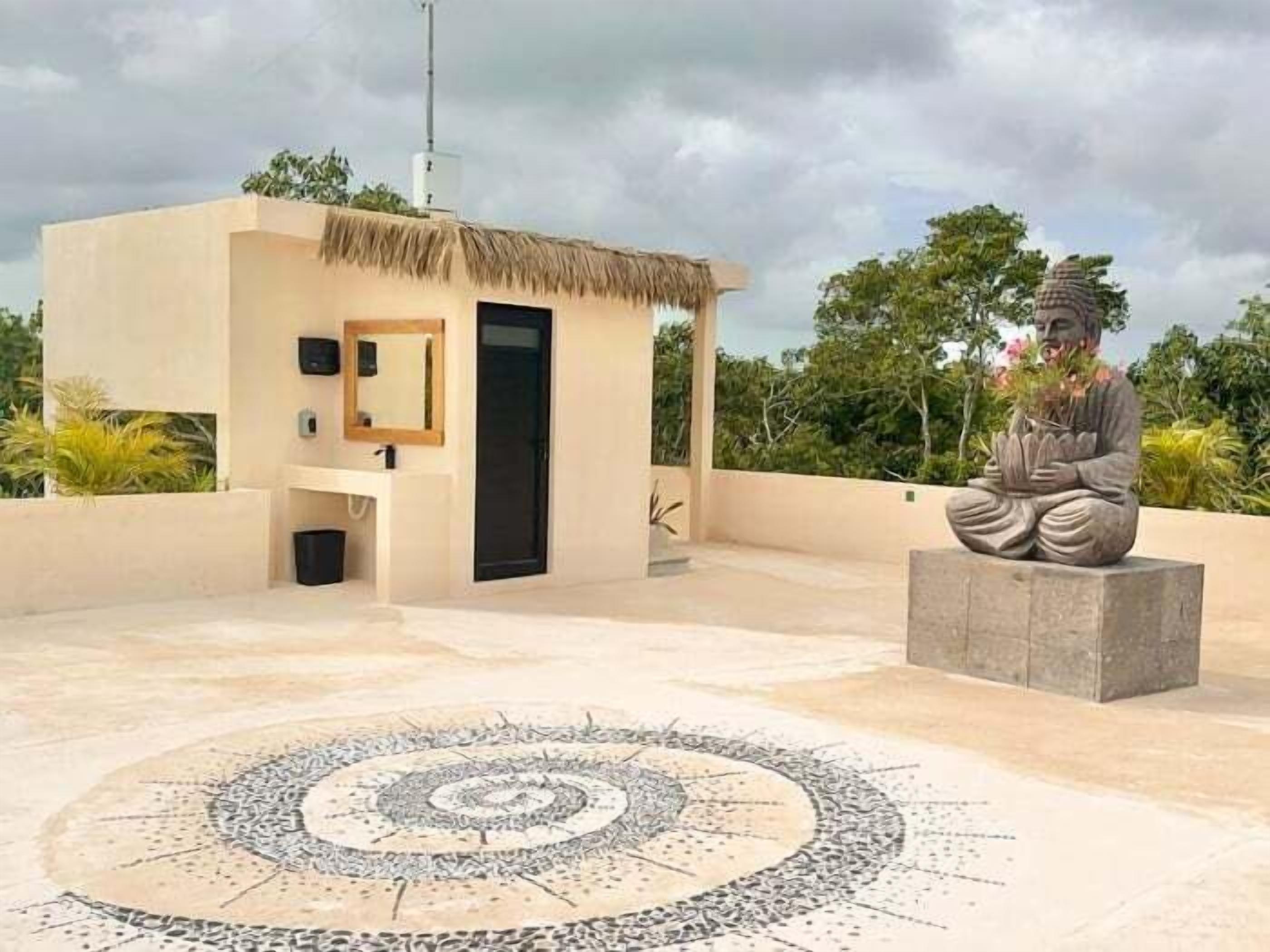 A modern outdoor bathroom with a thatched roof, a small sitting area with a statue, and a circular mosaic on the ground.