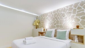 A modern hotel room with a stone wall, a neatly made bed with white linens, and a bedside table with a vase and a lamp.