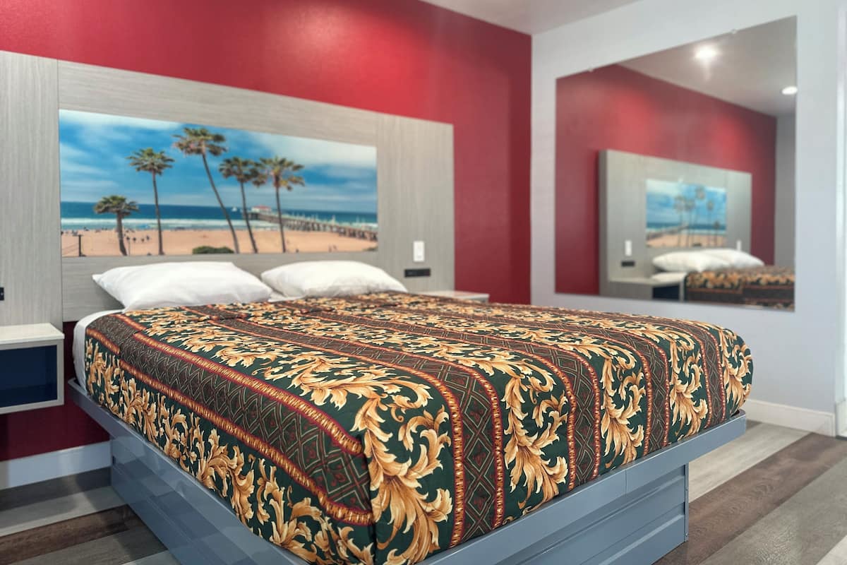 A bedroom with a bed, a nightstand, a large painting of a beach scene, and a mirror reflecting the bed.