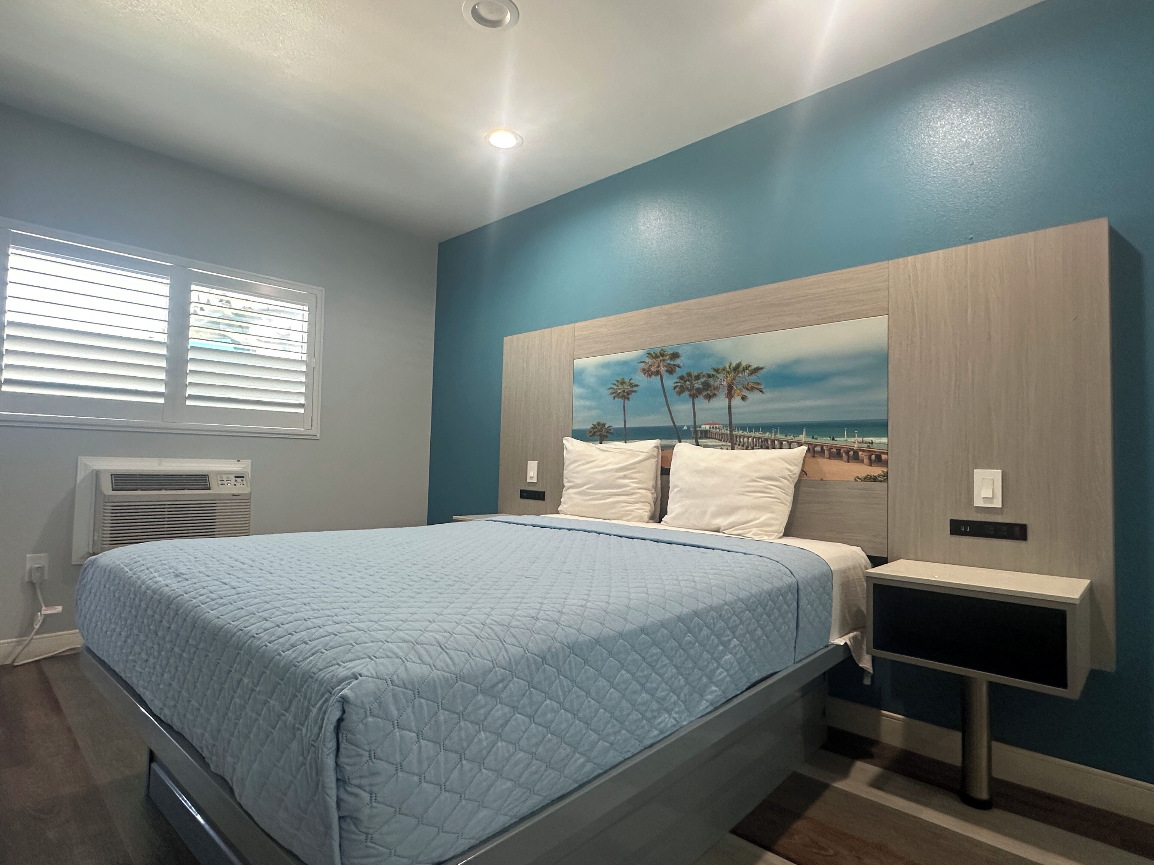 A bedroom with a bed, a nightstand, a wall-mounted air conditioner, and a window with blinds.