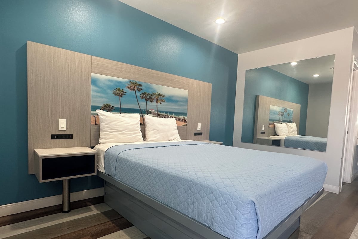 A modern bedroom with a large bed, a nightstand, a wall-mounted control panel, and a beach-themed artwork above the bed.