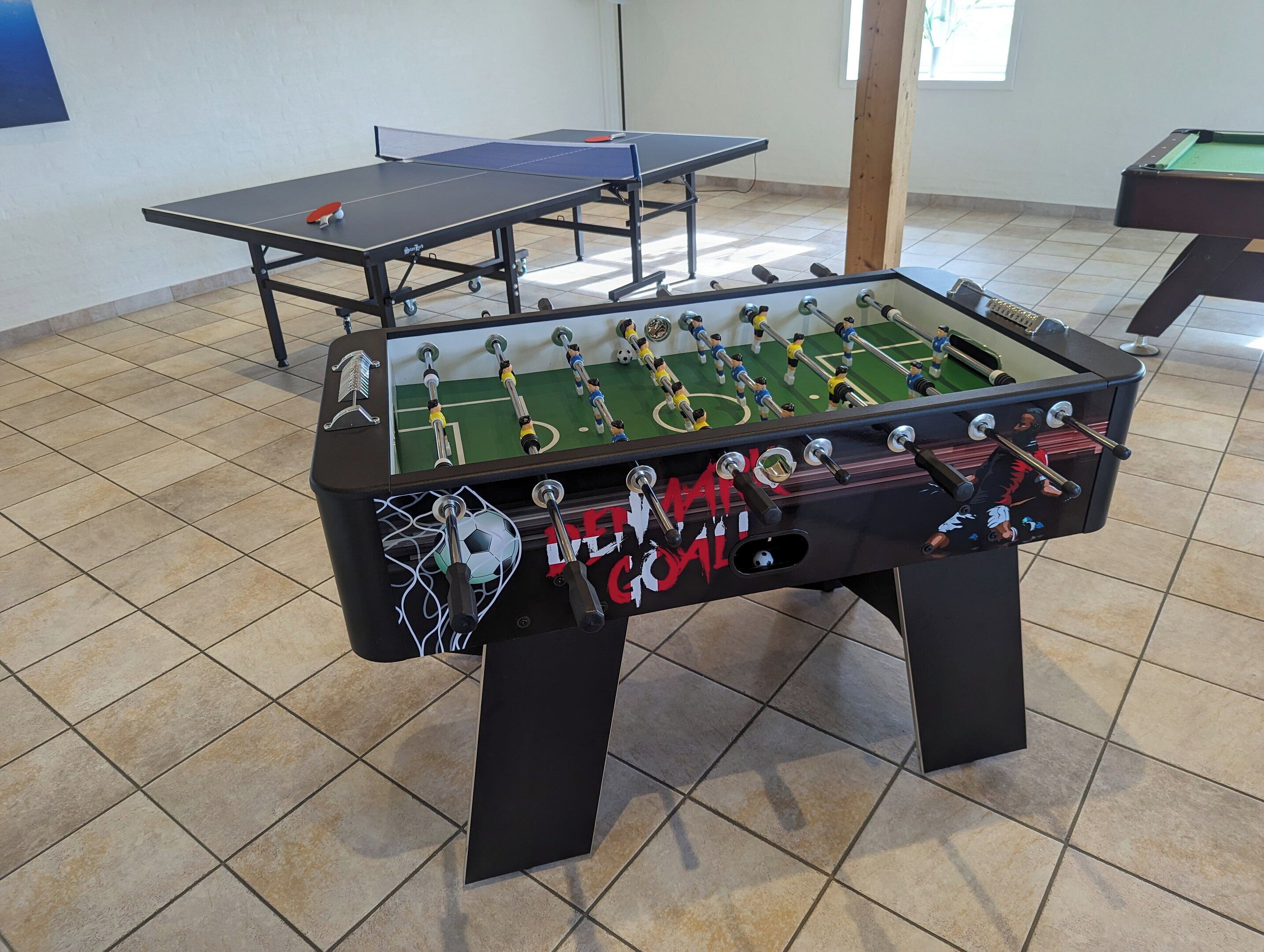 Une table de baby-foot avec une zone de but protégée par un filet, et une table de ping-pong en arrière-plan.