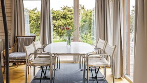 A room with a round table, four chairs, a vase with flowers, and a view of greenery through large windows. - Ringenäs by Interhome (Halmstad)