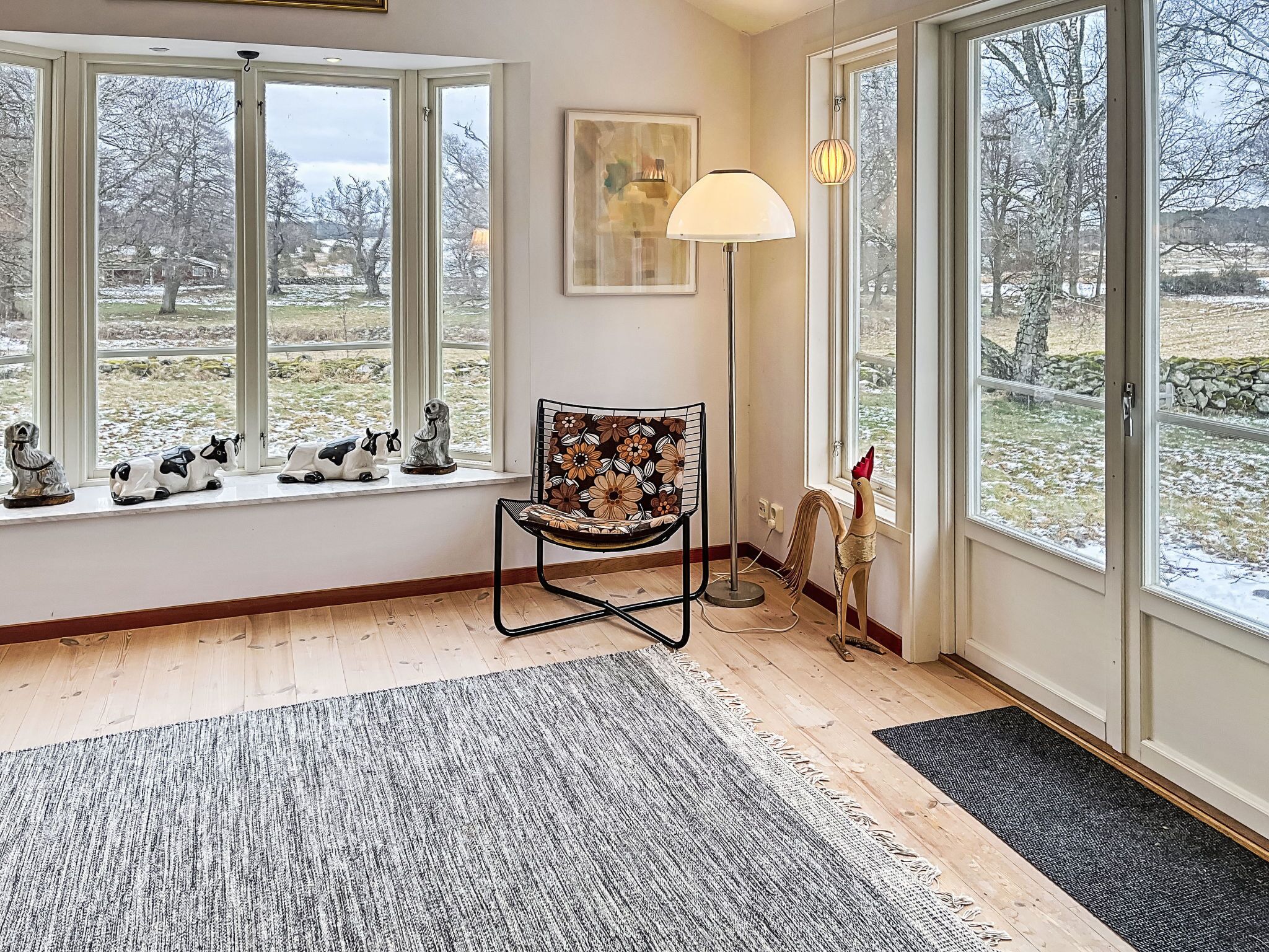 Ein Zimmer mit großen Fenstern, einem Sessel mit Blumenkissen, einer Stehlampe und einer Hahnschmuck.