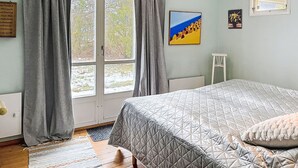 Ett sovrum med en stor säng, grå gardiner, ett fönster med utsikt över träd, en inramad tavla på väggen och ett litet bord.