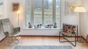 Ett rum med ett stort fönster, två stolar, en matta och olika inramade konstverk.