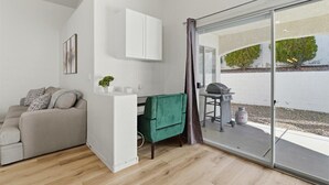 Een gezellig compact leefruimte met een bank, een klein tafeltje, een kast en een schuifdeur naar een buitenruimte met een barbecue.'