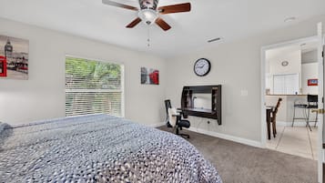 Ein Schlafzimmer mit Bett, Schreibtisch, Stuhl, Deckenventilator, Fenster mit Jalousien und einer Wanduhr.