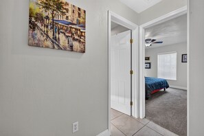 Un couloir orné d’un tableau représentant une scène de rue, une porte blanche donnant sur une chambre à coucher avec un lit et un ventilateur de plafond, et un sol recouvert d’une moquette.