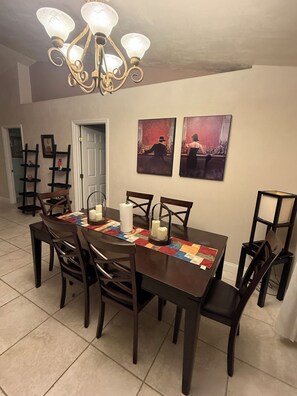 Une salle à manger avec une table en bois dressée pour six personnes, entourée de chaises. Un lustre se trouve au-dessus de la table, et deux œuvres d’art encadrées ornent le mur.