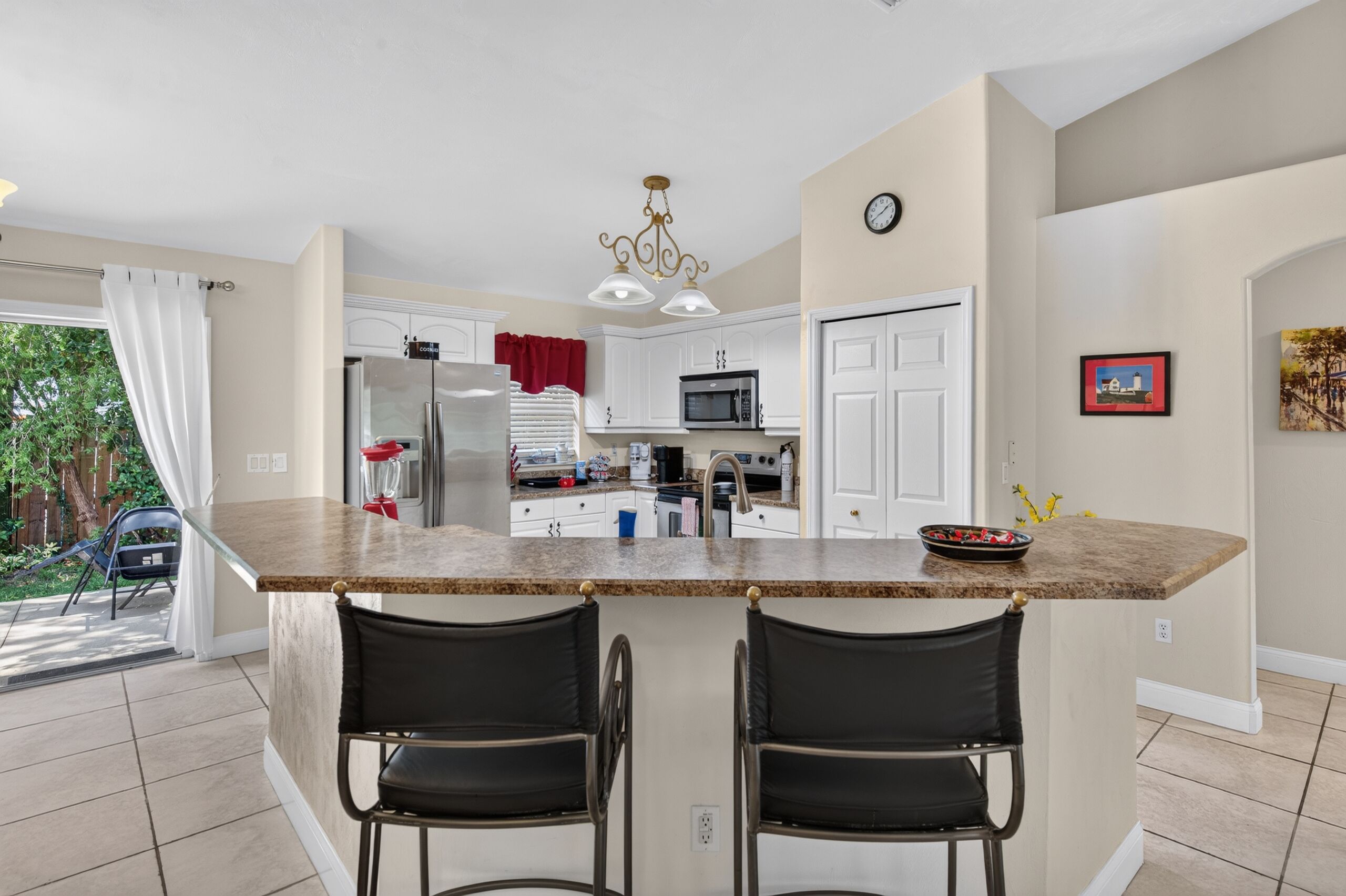 A kitchen with a central island, two bar stools, a refrigerator, a microwave, and a clock on the wall.