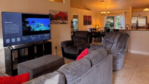 A living room with a flat-screen TV, grey sofas, and a dining area with a chandelier.