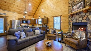 Een gezellig interieur van een chalet met een stenen open haard, houten wanden, een keuken en een zithoek met een bank, fauteuils en een salontafel.