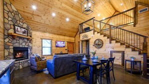 Een gezellig interieur van een chalet met een stenen open haard, een zithoek met een bank en een eettafel, en een trap naar een verdieping.