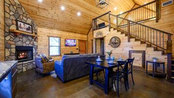 A cozy cabin interior with a stone fireplace, a living area with a sofa and a dining table, and a staircase leading to an upper level.