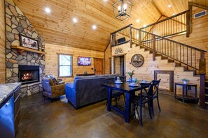 Ein gemütliches Blockhäuschen mit Kamin, einem Wohnbereich mit Sofa und Esstisch sowie einer Treppe zu einer oberen Ebene.