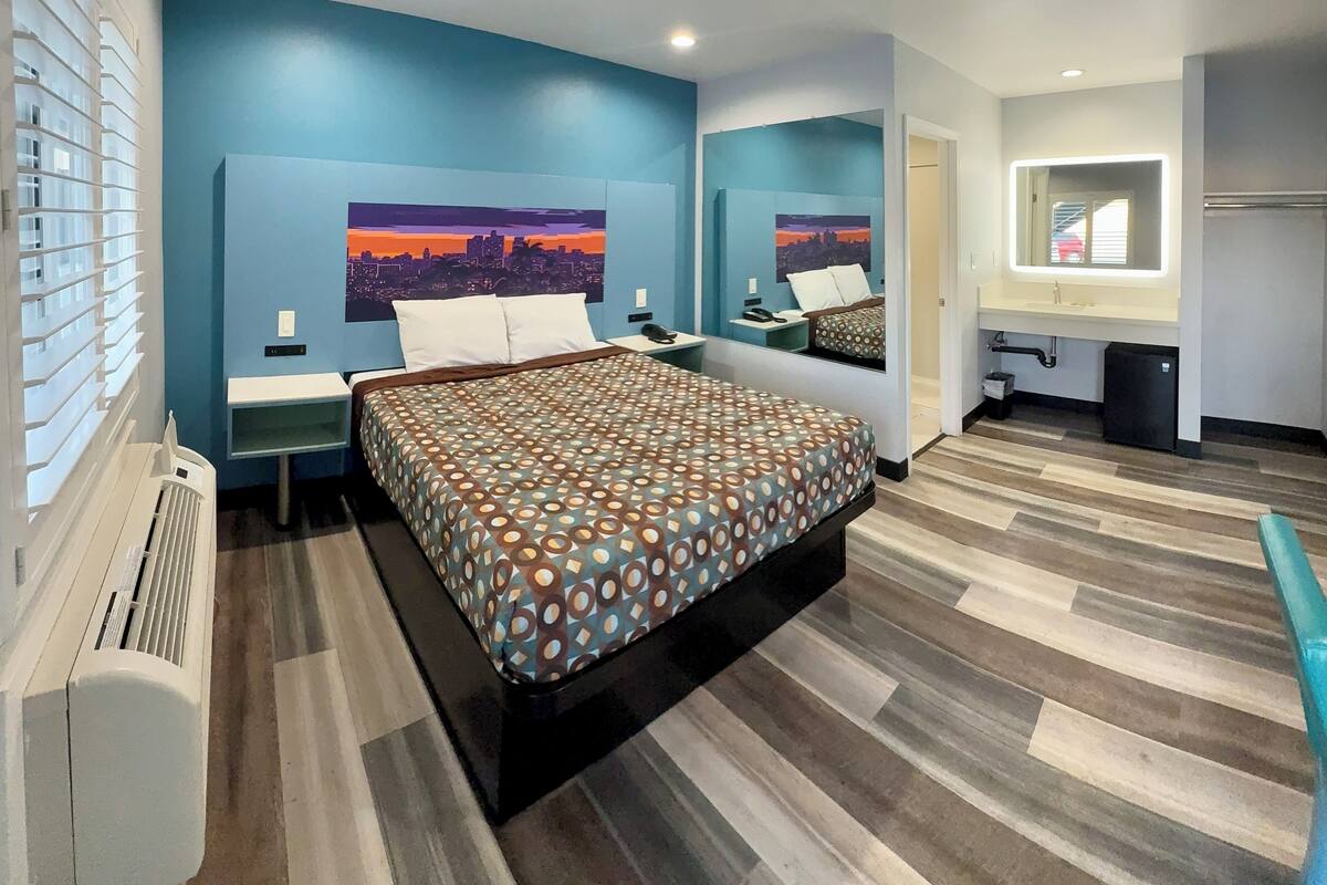 A modern hotel room with a large bed, a nightstand, a desk with a chair, a mirror, and a window with blinds.