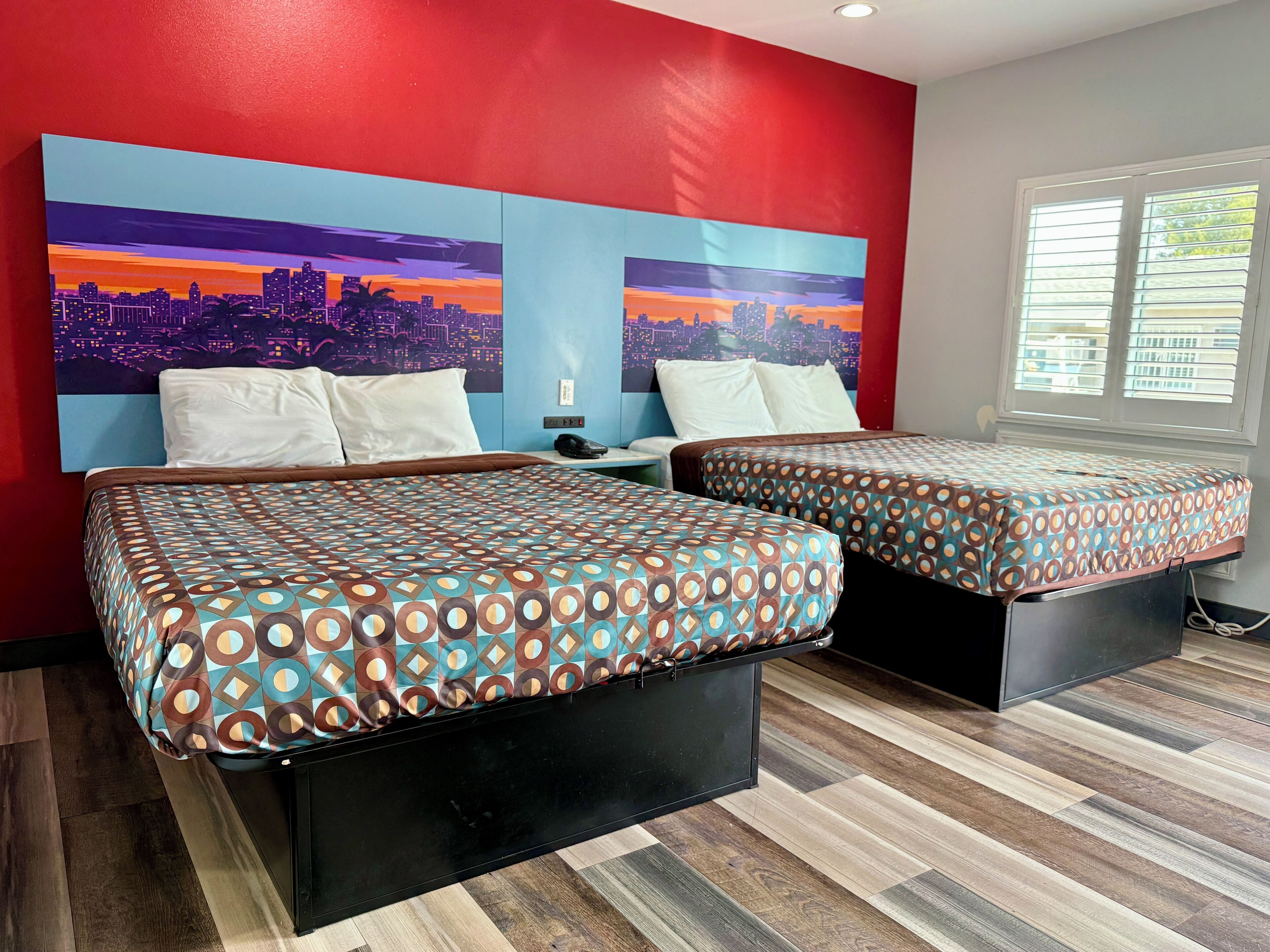 A hotel room with two beds, a cityscape mural on the wall, and a window with blinds.