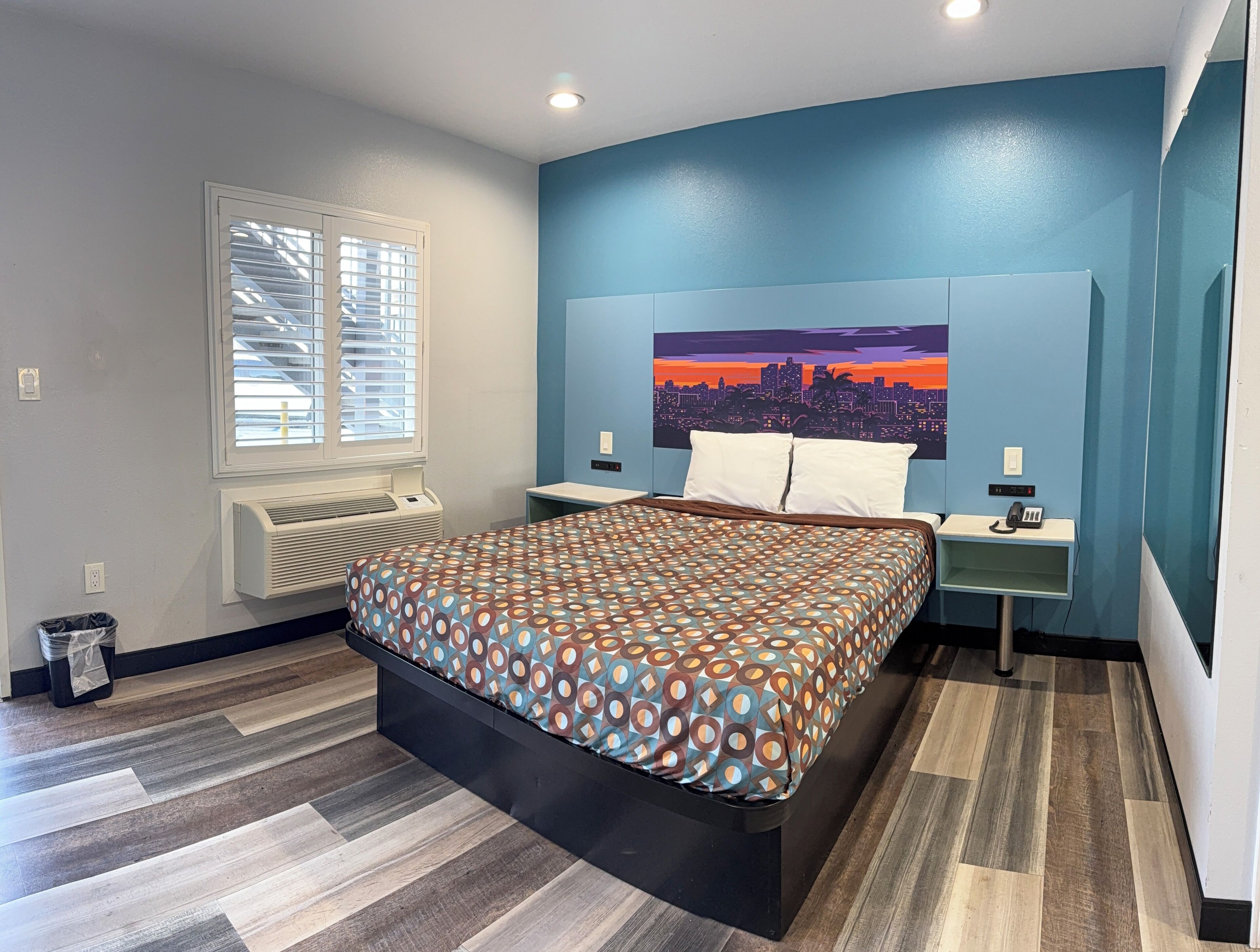 A bedroom with a bed, a nightstand, a wall-mounted phone, a window with blinds, and a wall-mounted air conditioner.